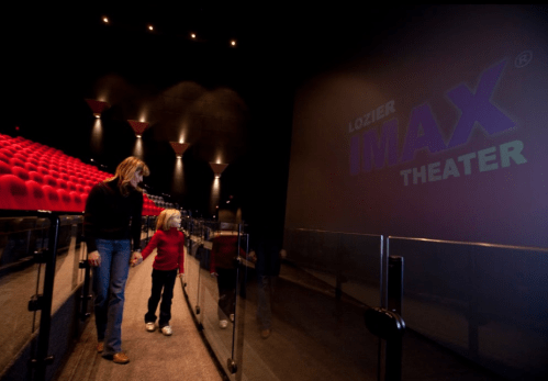 A woman and a child walk hand-in-hand in an IMAX theater with red seating and a large screen.