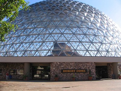 A large geodesic dome structure with a stone base, labeled "DESERT DOME," set against a clear blue sky.