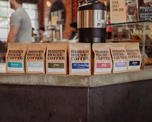A display of coffee bags labeled "Hidden House Coffee" in a café setting, with a coffee pot in the background.