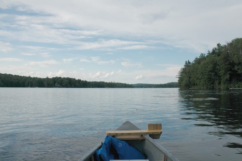 A calm lake scene viewed from a canoe, surrounded by trees under a cloudy sky.