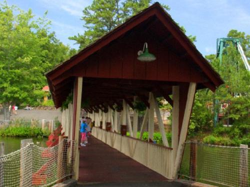 A covered wooden bridge with a red roof, surrounded by greenery and a calm waterway. Two people walk along the path.