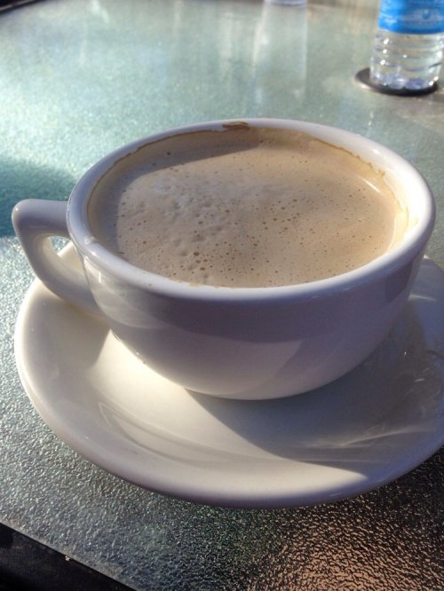 A white coffee cup filled with frothy coffee, resting on a saucer on a glass table.
