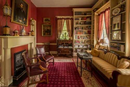 Cozy room with red walls, bookshelves, vintage furniture, and framed portraits, creating a warm, inviting atmosphere.