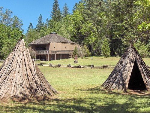 Two wooden structures resembling teepees in a grassy area, with a large building and trees in the background.