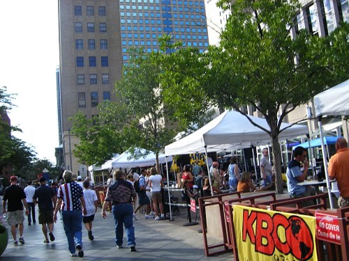 A bustling street scene with people walking past vendor tents and colorful banners in an urban setting.