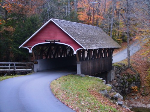 A red covered bridge spans a small stream, surrounded by autumn foliage and a winding road.