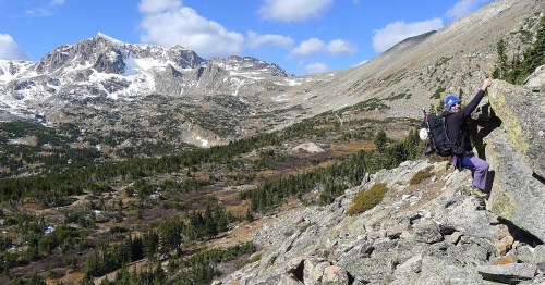 A climber ascends a rocky slope with snow-capped mountains and a lush valley in the background under a blue sky.