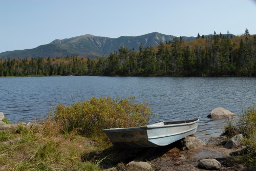 A small boat rests on the shore of a calm lake, surrounded by trees and mountains under a clear blue sky.
