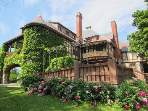 A large, ivy-covered mansion with a turret, surrounded by colorful flowers and greenery under a blue sky.