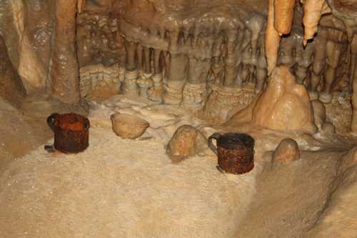 Rusty metal cups and a bowl sit on a sandy cave floor, surrounded by stalactites and stalagmites.