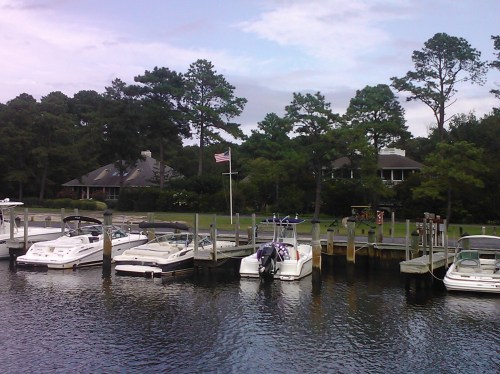 A peaceful marina with several boats docked, surrounded by trees and a house in the background, under a cloudy sky.