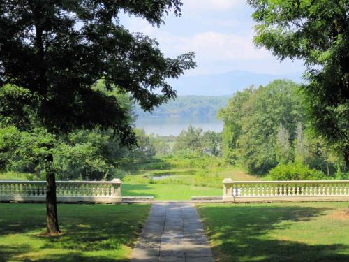 A serene landscape view featuring a pathway leading to a lake, framed by trees and distant mountains under a blue sky.