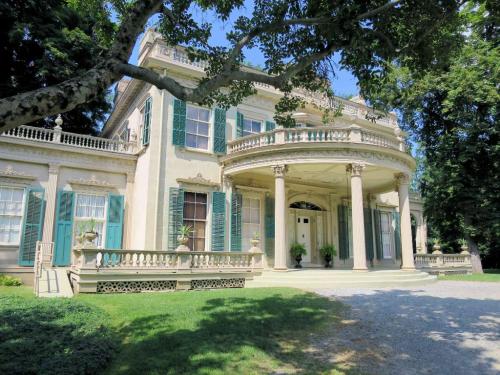 A grand historic mansion with green shutters, a large porch, and lush landscaping under a clear blue sky.