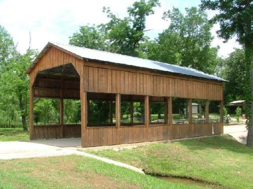 A wooden covered structure with a metal roof, surrounded by trees and grass, near a small waterway.