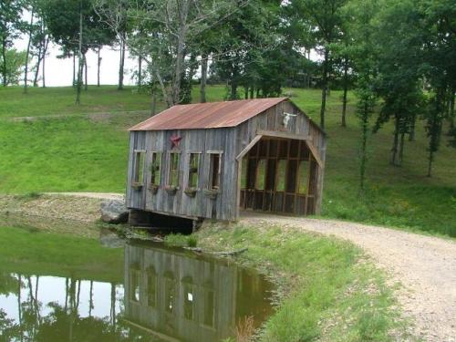A rustic wooden shed with a metal roof sits by a pond, surrounded by trees and a gravel path. Reflection in the water.
