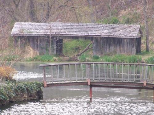 A weathered wooden structure beside a calm pond, with a small bridge in the foreground and greenery surrounding the scene.