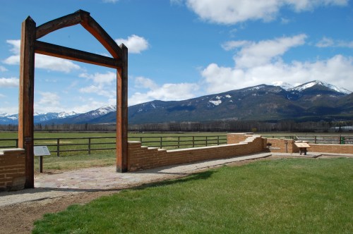 A wooden archway stands in a grassy area with mountains in the background under a partly cloudy sky.