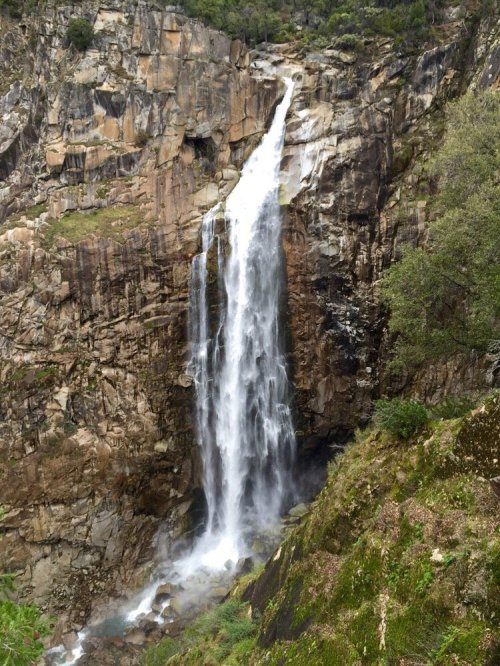 A tall waterfall cascading down rocky cliffs, surrounded by lush greenery and rugged terrain.