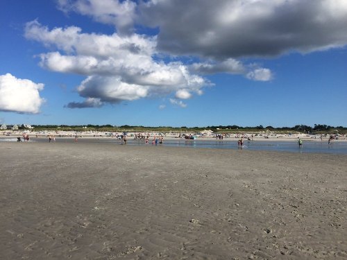A sandy beach with people enjoying the sun, scattered clouds, and a clear blue sky in the background.