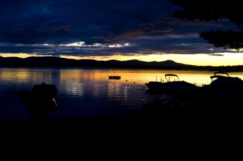 A serene lake at sunset, with boats silhouetted against a colorful sky and distant mountains.