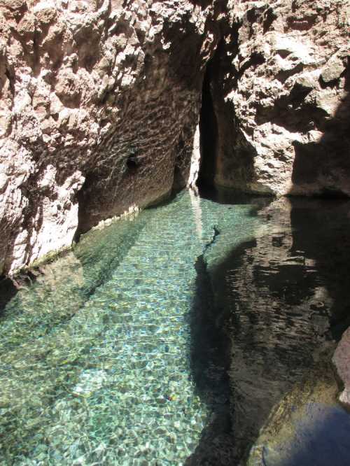 A serene, clear pool of water surrounded by rocky walls, reflecting sunlight in a natural setting.