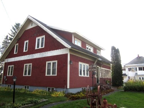 A red bed and breakfast building with a sign, surrounded by greenery and other structures in the background.