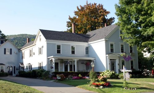 A charming two-story house with a landscaped yard and colorful flowers, surrounded by trees and hills in the background.