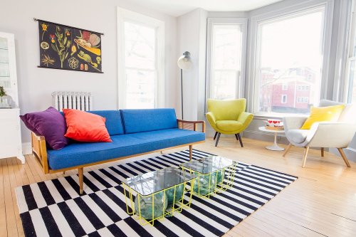 Bright living room with a blue sofa, colorful cushions, modern chairs, and striped rug, featuring large windows and natural light.