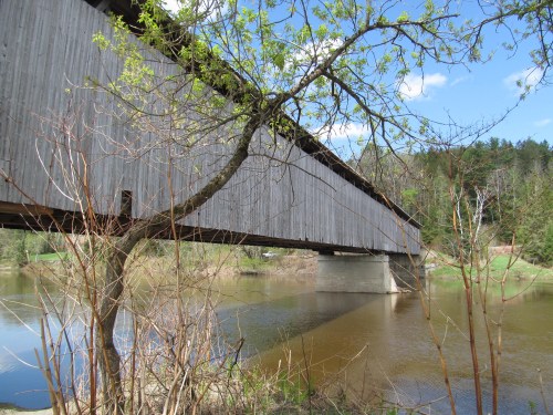 A wooden covered bridge spans a calm river, surrounded by trees and greenery under a clear blue sky.