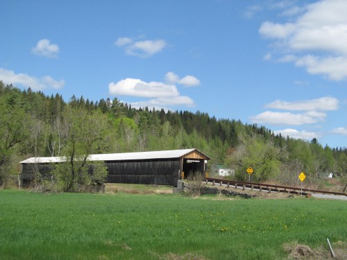 A covered wooden bridge spans a grassy field, surrounded by trees and a blue sky with fluffy clouds.
