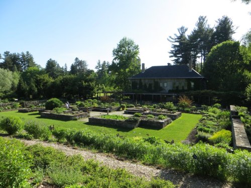 A lush garden with neatly arranged flower beds and a house in the background, surrounded by trees and greenery.