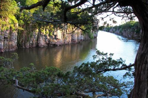 A serene river flows between rocky cliffs, framed by lush green trees under a clear sky.