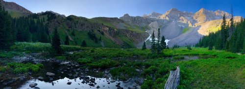 A panoramic view of a lush green valley with mountains, a stream, and trees under a clear blue sky.