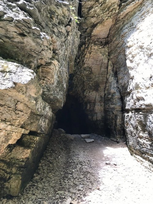 A narrow rocky canyon with steep walls and a dark opening at the end, surrounded by scattered stones on the ground.