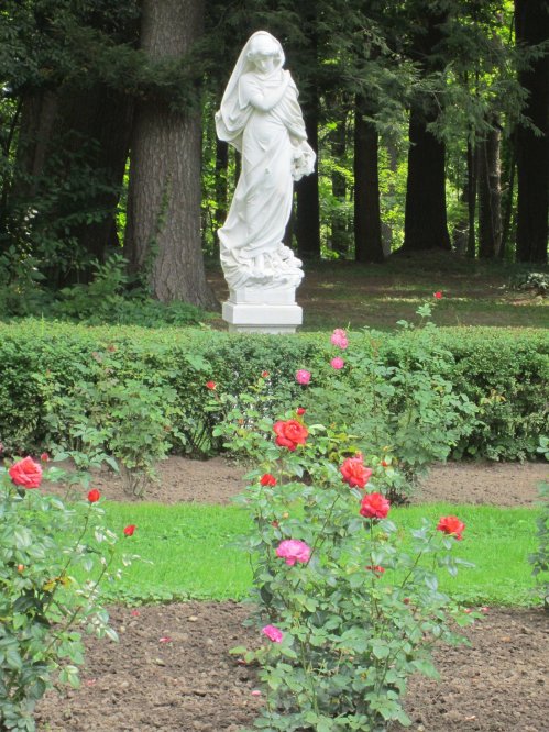 A white statue of a woman stands among blooming rose bushes in a lush garden surrounded by trees.