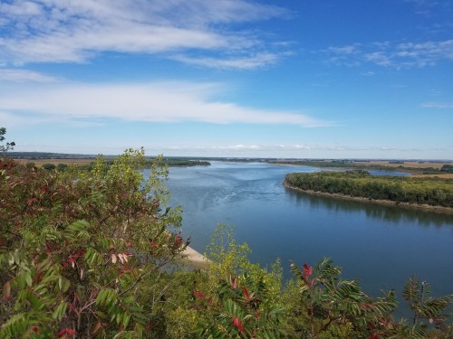 A serene river flows through lush greenery under a bright blue sky with scattered clouds.