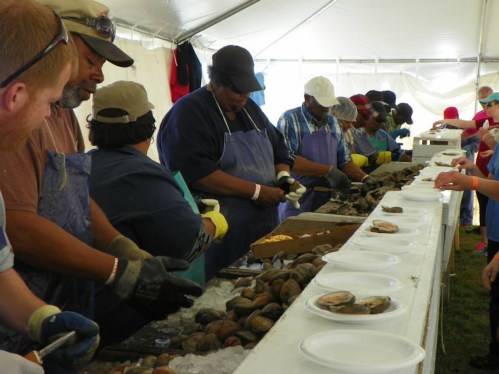 A group of people shucking oysters at a long table under a tent, with plates and seafood displayed.