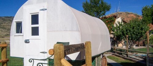 A white covered wagon parked near a wooden fence, surrounded by trees and hills in the background.