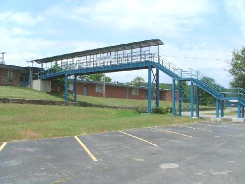 A blue metal walkway connects two buildings, set against a grassy area and cloudy sky. Parking spaces are visible below.