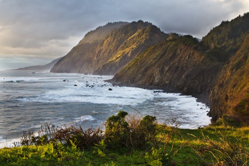 Coastal landscape with rugged cliffs, waves crashing on the shore, and lush greenery under a cloudy sky.