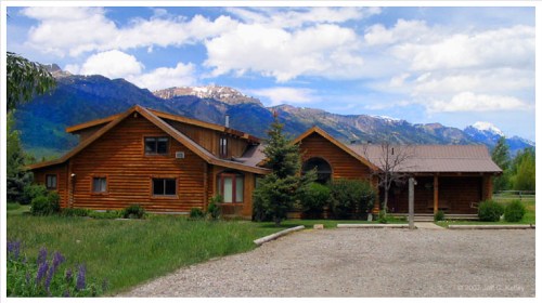 A rustic wooden house surrounded by greenery, with mountains and a blue sky in the background.