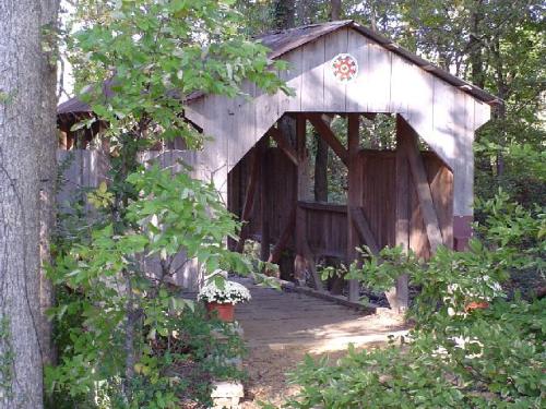 A rustic wooden covered bridge surrounded by trees and greenery, with a flower pot at the entrance.