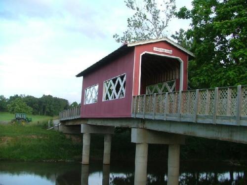 A red covered bridge with white trim spans a calm river, surrounded by greenery and a cloudy sky.