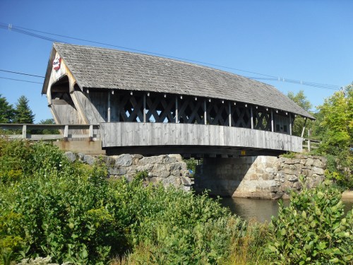 A wooden covered bridge spans a river, surrounded by greenery and a clear blue sky.