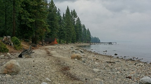 A rocky shoreline with trees and a pier under a cloudy sky, featuring a cabin in the background.