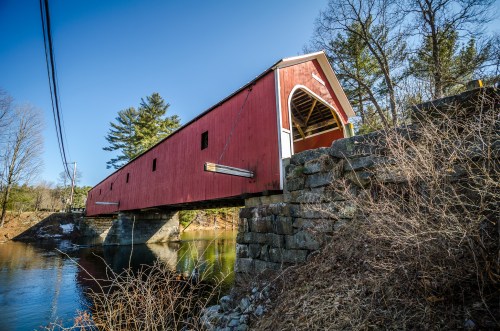 A red covered bridge spans a calm river, surrounded by trees and a clear blue sky.
