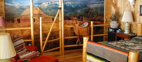 Cozy rustic bedroom with wooden decor, a large mountain mural, and a saddle on display. Warm lighting from lamps.