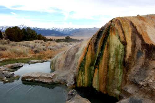 A natural hot spring with colorful mineral deposits, surrounded by mountains and grassy terrain under a cloudy sky.