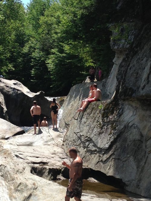 People enjoying a sunny day by a rocky river, some sitting on rocks and others wading in the water.