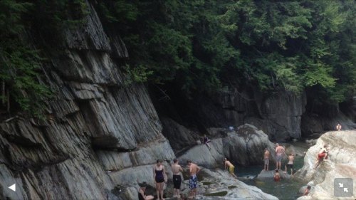 People swimming and relaxing in a rocky river surrounded by lush green trees.